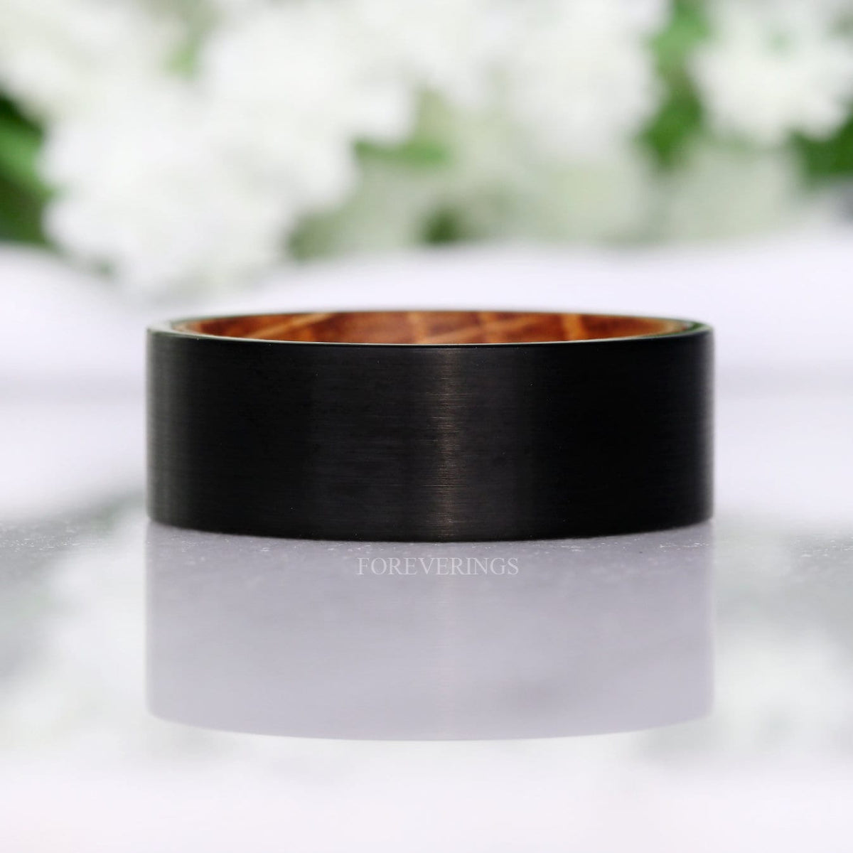 Black brushed tungsten band shown straight on, whiskey barrel wood visible at the rim