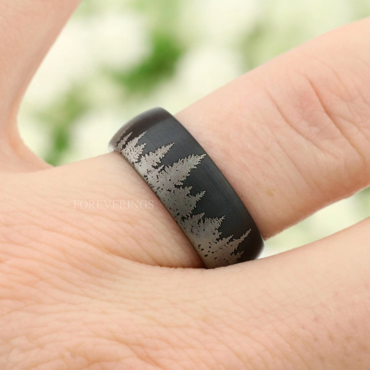 Men's black tungsten ring with etched pine tree line worn on a finger, angled from above