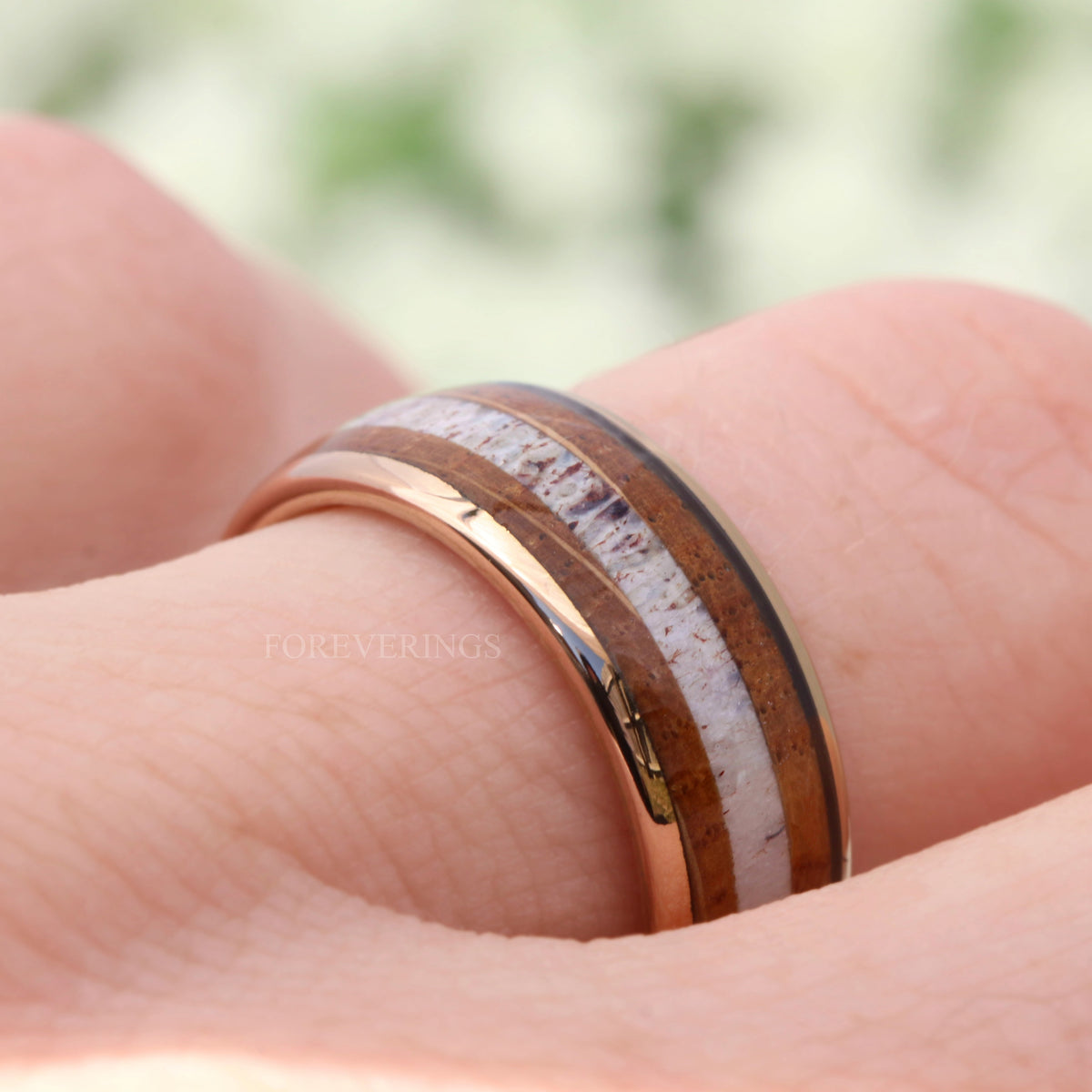 Men's rose gold tungsten band with whiskey barrel wood and antler inlay, worn on a finger
