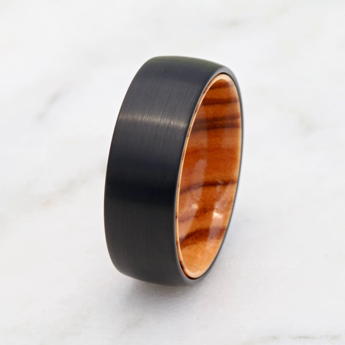 Black tungsten olive wood band standing on white marble, seen from farther back