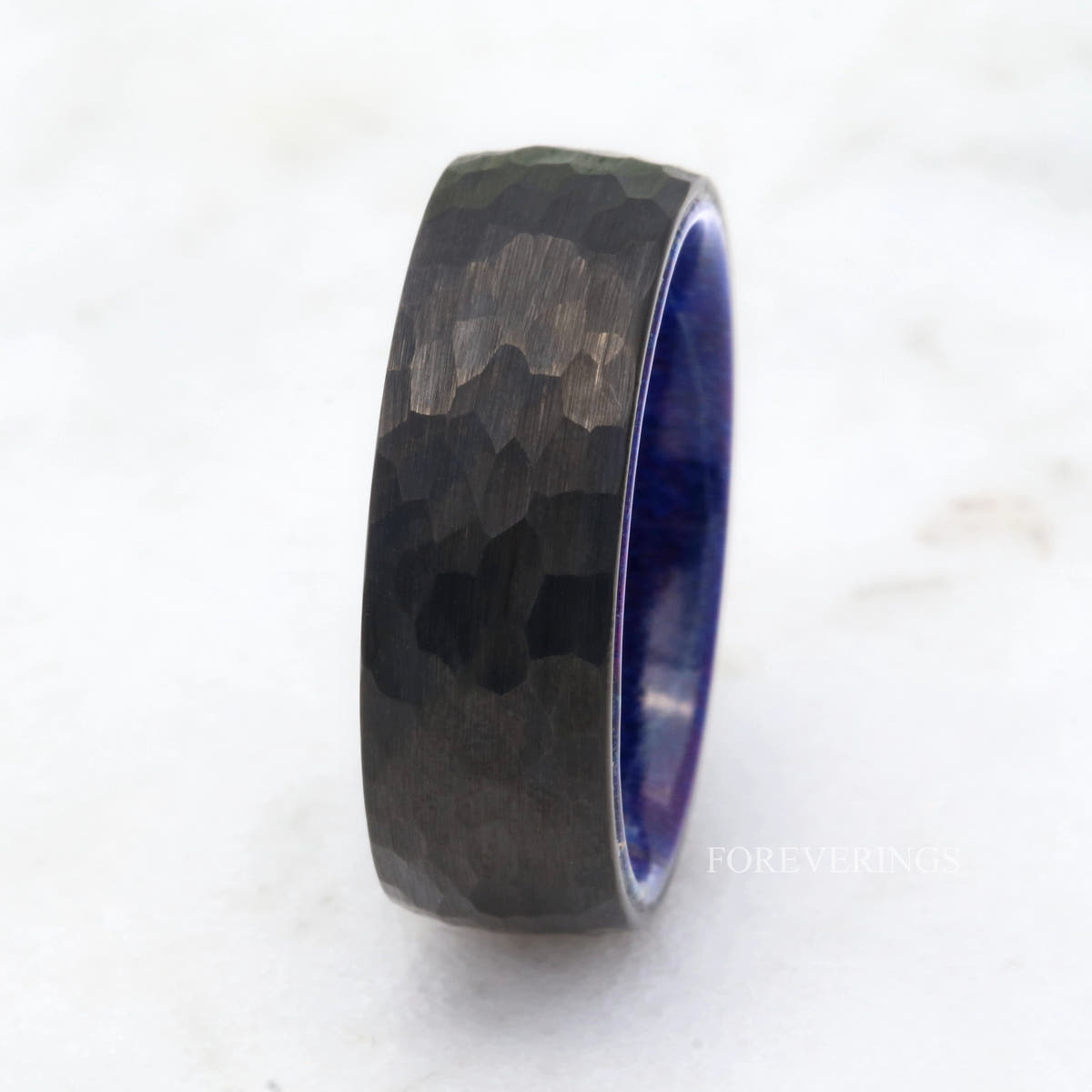 Hammered black tungsten band shown mostly side-on, with a sliver of the blue wood lining