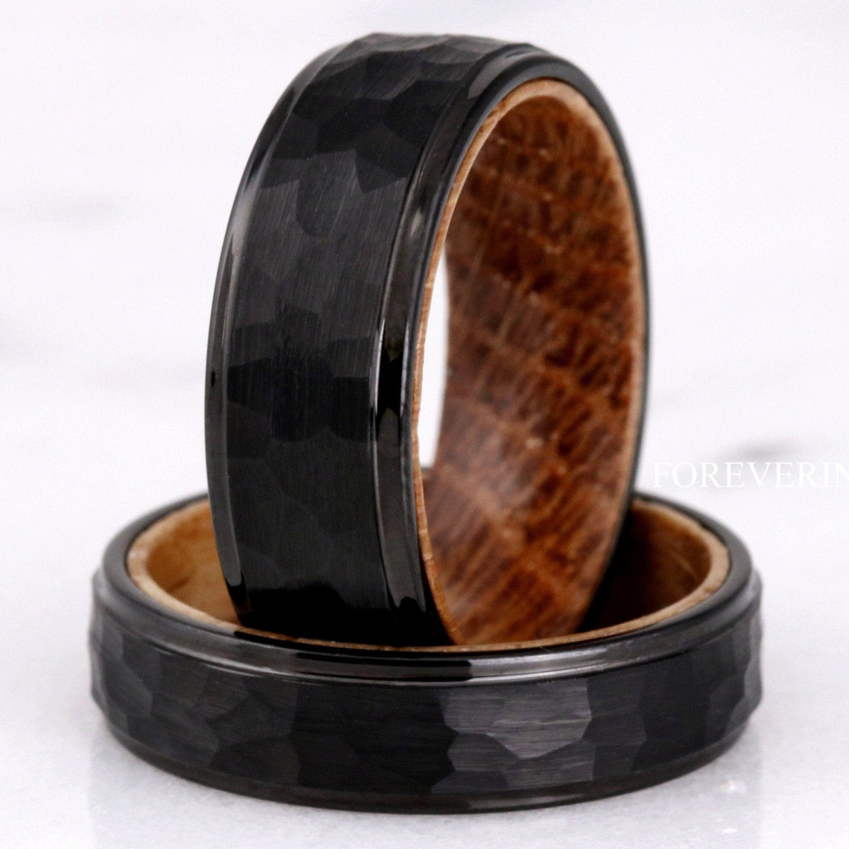 Men's hammered black tungsten bands lined with whiskey barrel wood, one standing on the other
