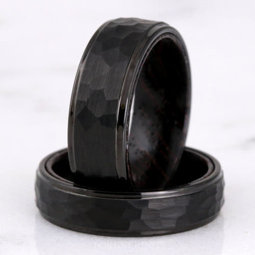 Men's hammered black tungsten band with wenge wood lining standing inside a second band lying flat