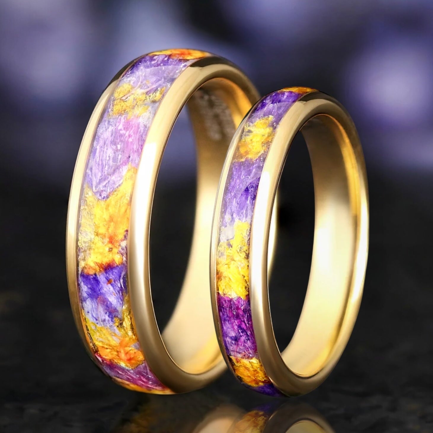Two gold rings with colorful inlay on a reflective surface with a blurred background