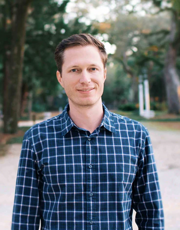 Smiling person in a blue plaid shirt standing outdoors under oak trees