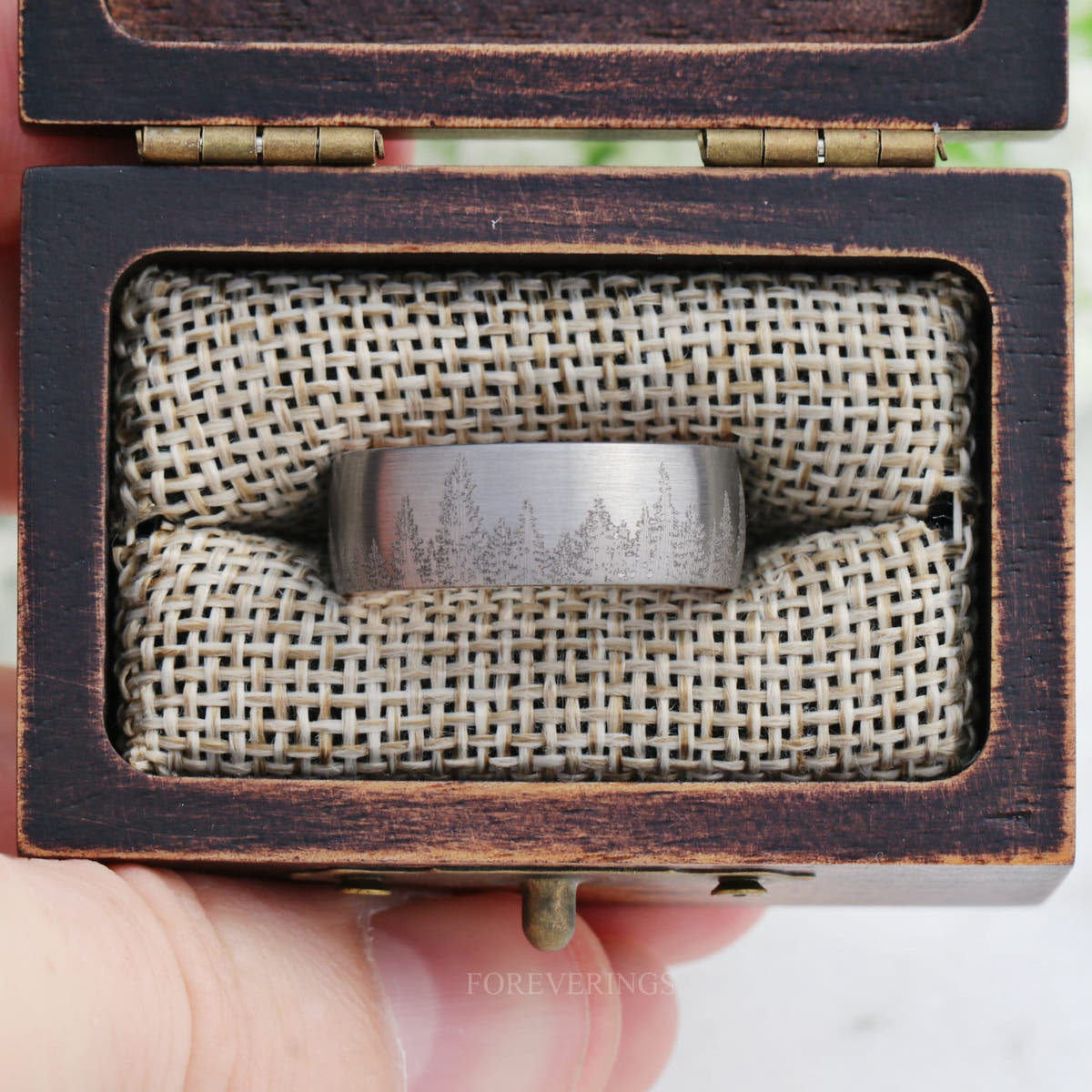 Wood ring box held open in a hand, showing a band seated in the burlap insert