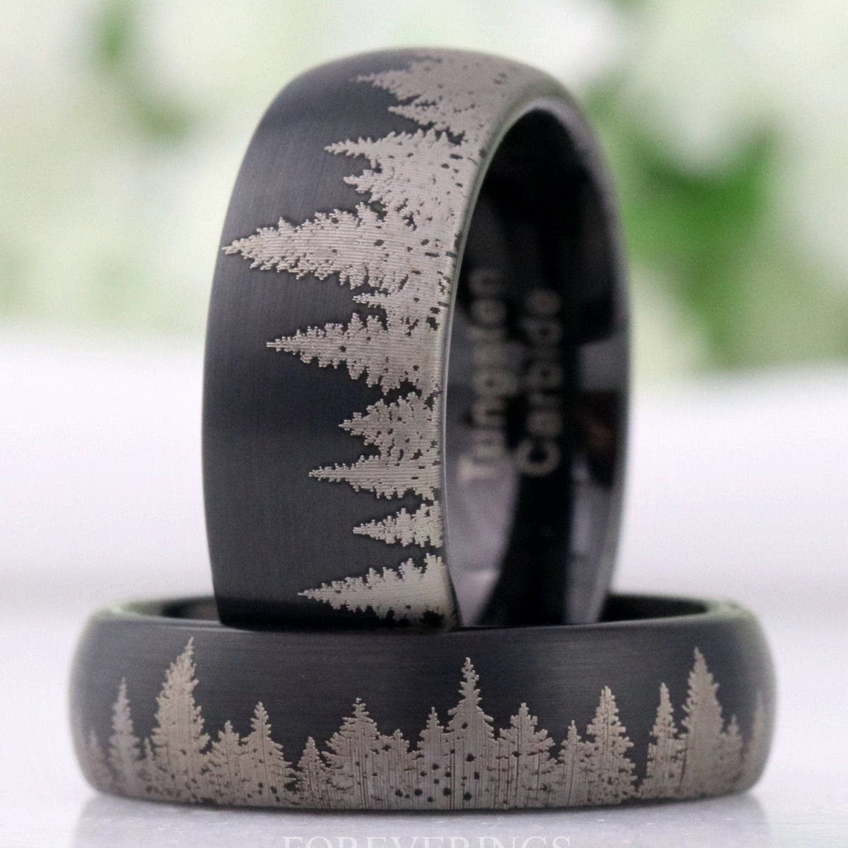 Men's matte black tungsten wedding bands with etched pine tree line, one standing on another