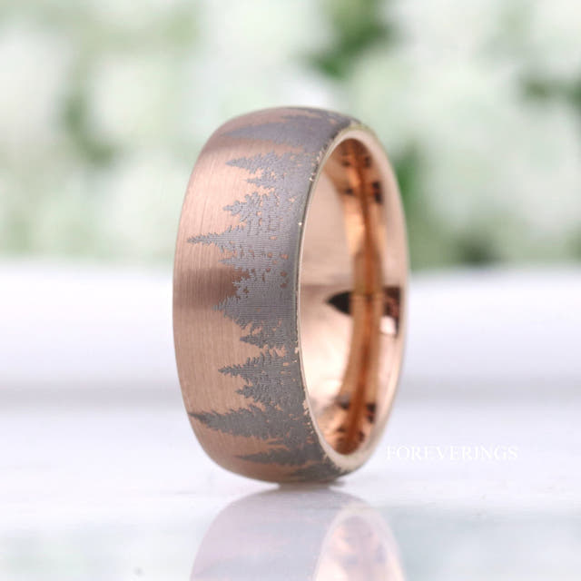 Matte rose gold tungsten band with an etched pine tree landscape, standing at an angle