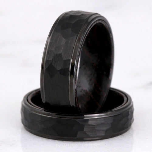 Men's hammered black tungsten band with wenge wood lining standing inside a second band lying flat