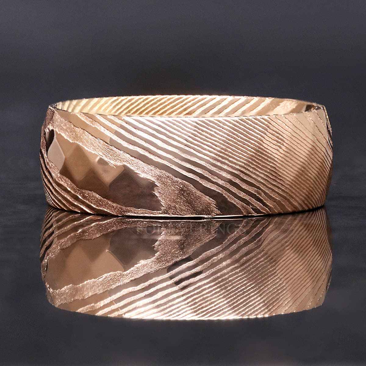 Men's 8mm rose gold Damascus steel ring with hammered facets, lying flat with reflection below