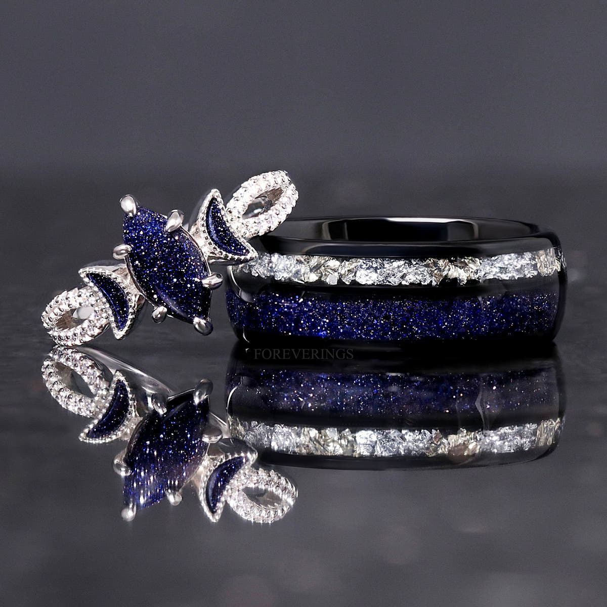 8mm black tungsten band beside a silver women's ring with a marquise blue sandstone center, reflection below